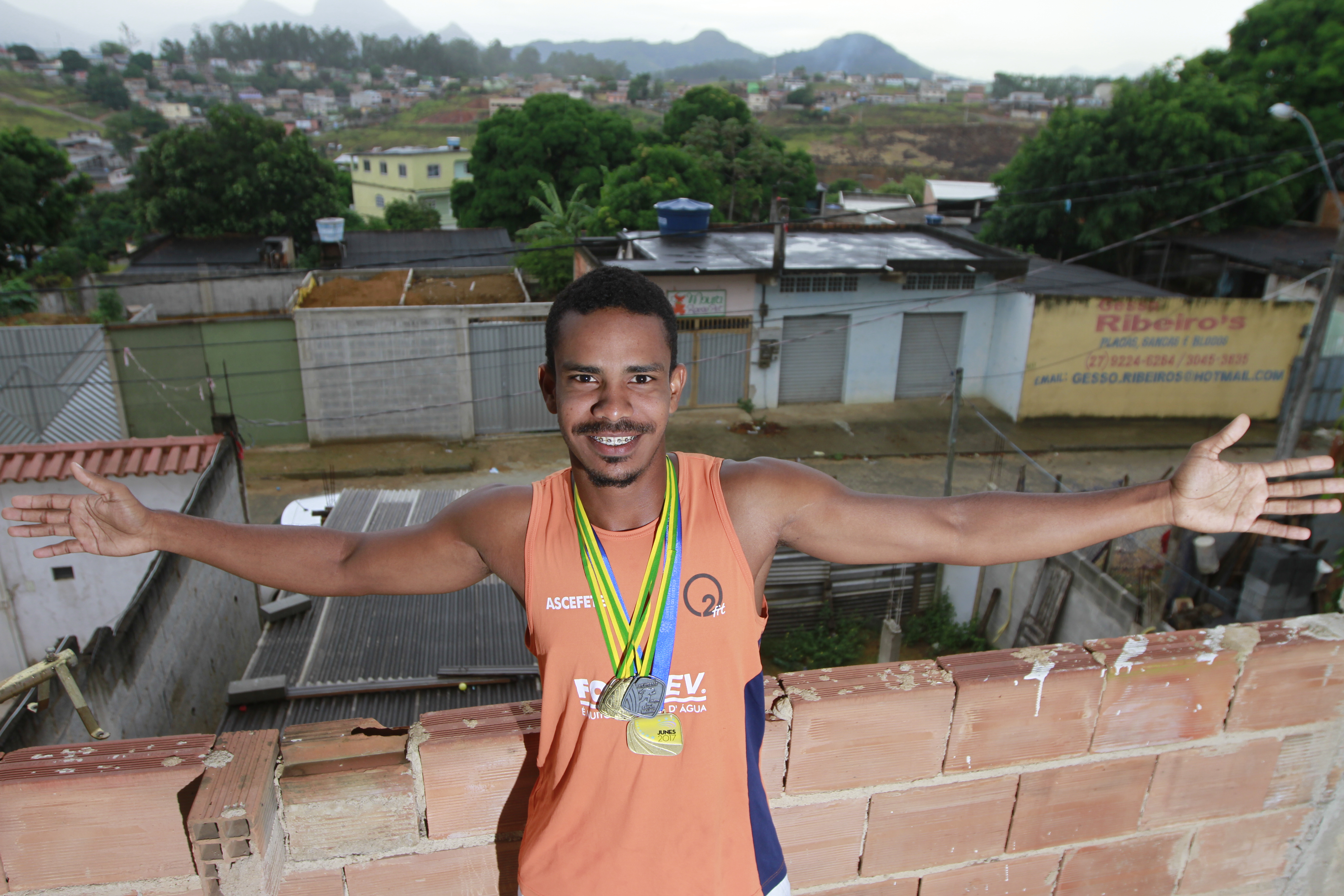 Eduardo Santos no terraço de sua casa, em Nova Bethania, Viana, abre os braços com a vista do bairro ao fundo. Foto: Rodrigo Gavini/AT