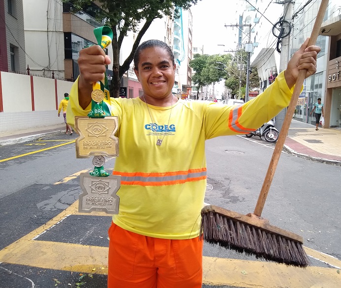 Juliana segura sua medalha e a vassoura que utiliza todos os dias para fazer a limpeza das ruas em Guarapari (Foto: Roberta Bourguignon)
